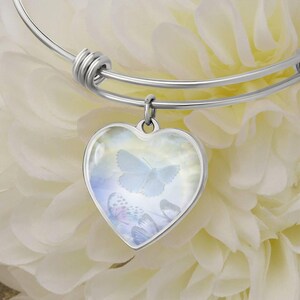 May include: A silver bangle bracelet with a heart-shaped charm featuring a delicate blue butterfly design. The charm is surrounded by a white background with a subtle cloud-like pattern.