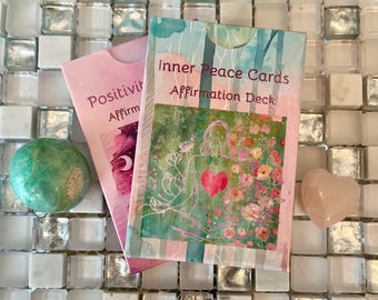 May include: Two pink and white card decks with the text "Inner Peace Cards Affirmation Deck" and "Positivi Affirm" on the front. The decks are sitting on a white tiled surface with a green and white stone and a pink heart-shaped stone.