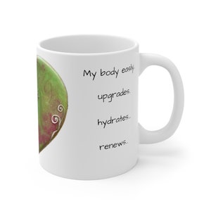 May include: White ceramic mug with a green and red heart-shaped design on the side. The mug has the text "My body easily upgrades, hydrates, renews..." in black. The mug has a curved handle.