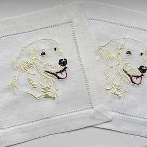 May include: Two white linen cocktail napkins with embroidered images of a golden retriever dog. The dog is facing right and has its tongue sticking out.