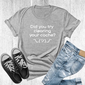 Pode incluir: Camiseta cinza com o texto "Did you try clearing your cache?" e um emoticon japonês. A camiseta está colocada sobre uma superfície de madeira com um par de tênis pretos e uma calça jeans azul.