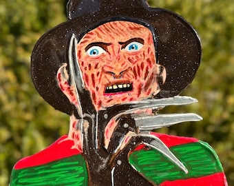 May include: A resin wall hanging featuring a stylized depiction of Freddy Krueger. The artwork showcases a brown hat, a scarred face with blue eyes, and metal claws. The design incorporates red and green stripes on the clothing, with a chain for hanging.