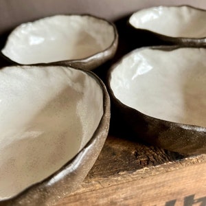 May include: Four small white ceramic bowls with a dark brown rim. The bowls are sitting on a wooden surface.