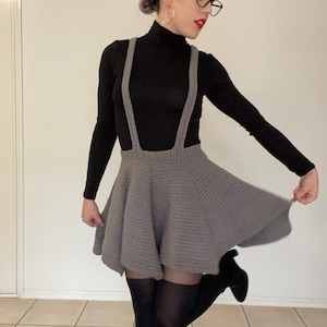 Op de afbeelding: Een grijze gehaakte rok met bretels, gecombineerd met een zwarte top met lange mouwen. De rok heeft een uitlopend ontwerp. De outfit wordt gecompleteerd met zwarte panty's en enkellaarsjes. De achtergrond is een effen muur en een tegelvloer.