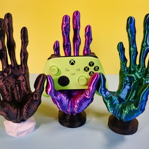 May include: Three 3D printed hands holding a green and black Xbox controller. The hands are purple, black, and blue and green.