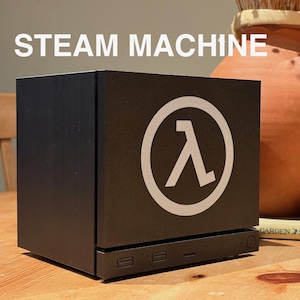 Puede incluir: Una Steam Machine negra con un logotipo blanco. El texto "STEAM MACHINE" está encima de la máquina. Un jarrón de terracota y un libro están en el fondo. Diseño moderno.