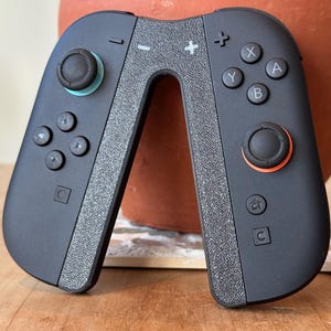 以下が含まれることがあります： 中央にテクスチャ加工されたグレーのストライプが入った黒いゲームコントローラー。コントローラーには黒いボタンがあり、左側にターコイズ色のジョイスティック、右側にオレンジ色のジョイスティックがあります。コントローラーはゲーム用に設計されています。