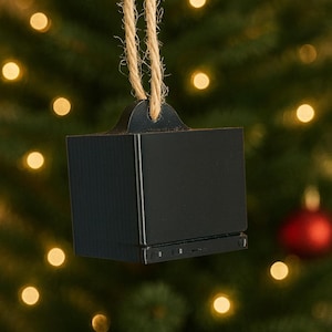 Könnte beinhalten: Ein schwarzer, würfelförmiger Weihnachtsschmuck, der einem alten Fernseher ähnelt. Er hängt an einer Schnur aus Naturfaser vor einem verschwommenen Hintergrund eines Weihnachtsbaums mit goldenen Lichtern und rotem Schmuck.
