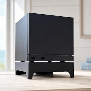 May include: A matte black, cube-shaped electronic device resting on a matching black stand with four short legs. The front panel features two USB ports, a small horizontal slot, and a circular power button located on the lower section of the unit.