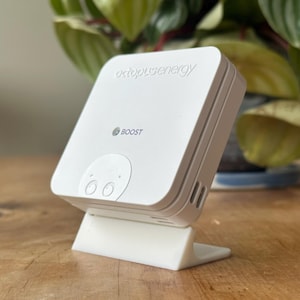 May include: A white electronic device with the text "octopusenergy" and "BOOST" printed on it. The device is mounted on a white stand. The background includes a green plant and a wooden surface.