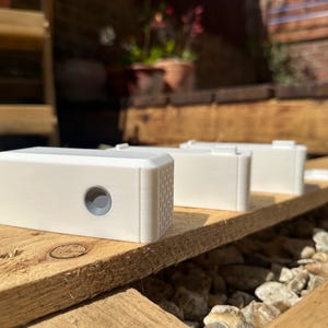 May include: Three white rectangular devices with a small round button on the front, sitting on a wooden surface.