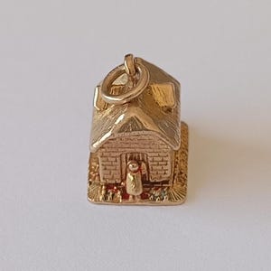 May include: A gold charm in the shape of a small house with a textured roof, brick-patterned walls, and a figure standing in the doorway. The charm features a circular jump ring at the top for attachment and rests on a square base.