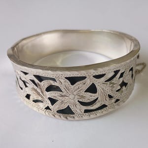 May include: A silver hinged bangle bracelet with a detailed floral and leaf design. The bracelet has a cut-out pattern, showing a dark interior. The metal has a polished finish, and the bracelet is approximately 6.35 cm in diameter.