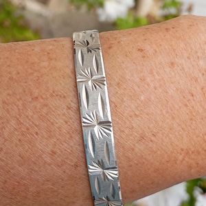 Può includere: Un bracciale rigido in metallo color argento caratterizzato da un motivo inciso ripetitivo di stelle e forme ovali allungate. La superficie del bracciale ha una finitura spazzolata e il design è inciso nel metallo per tutta la lunghezza visibile.