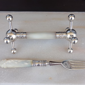 Puede incluir: Un reposacuchillos plateado y un tenedor. El reposacuchillos tiene una sección central blanca con bandas plateadas decorativas y cuatro brazos plateados que terminan en esferas. El tenedor tiene un mango blanco y detalles plateados.