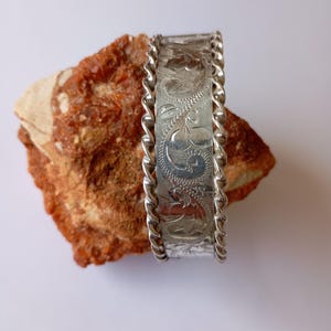 May include: A silver cuff bracelet with an engraved floral design. The bracelet has a twisted rope border on both sides. The cuff is resting on a rough, reddish-brown stone.