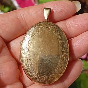Può includere: Un ciondolo a medaglione ovale color oro caratterizzato da un pannello centrale liscio e spazzolato, circondato da un intricato motivo a bordo inciso. Il medaglione include una piccola contromaglia lucida sulla parte superiore per una catenina.