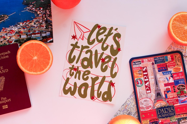 May include: A travel-themed still life featuring a postcard with the text "Let's travel the world", a passport, a Paris souvenir tray, and orange slices. The postcard has a red paper airplane and stars.