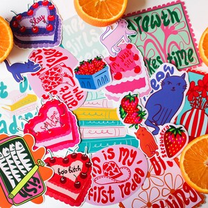 Puede incluir: Surtido de pegatinas coloridas con varios dise&ntilde;os, que incluyen un pastel en forma de coraz&oacute;n con la palabra "Slay" en &eacute;l, un gato con el texto "Just a girl", un jarr&oacute;n con flores y una lata de sardinas con el texto "Shut I'm readin".