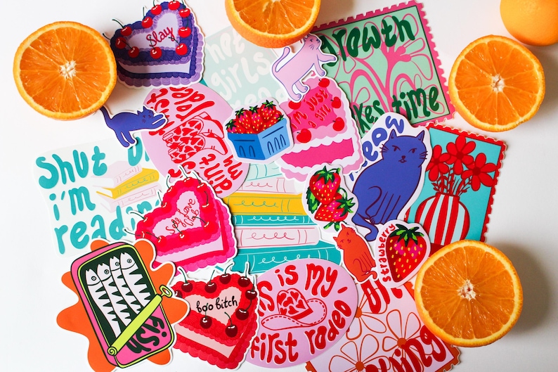 Puede incluir: Surtido de pegatinas coloridas con varios dise&ntilde;os, que incluyen un pastel en forma de coraz&oacute;n con la palabra "Slay" en &eacute;l, un gato con el texto "Just a girl", un jarr&oacute;n con flores y una lata de sardinas con el texto "Shut I'm readin".