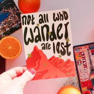 May include: A travel-themed image featuring a postcard with the text "not all who wander are lost" and a mountain illustration. Other items include a passport, a Paris souvenir, and orange slices. The postcard is held by a person.
