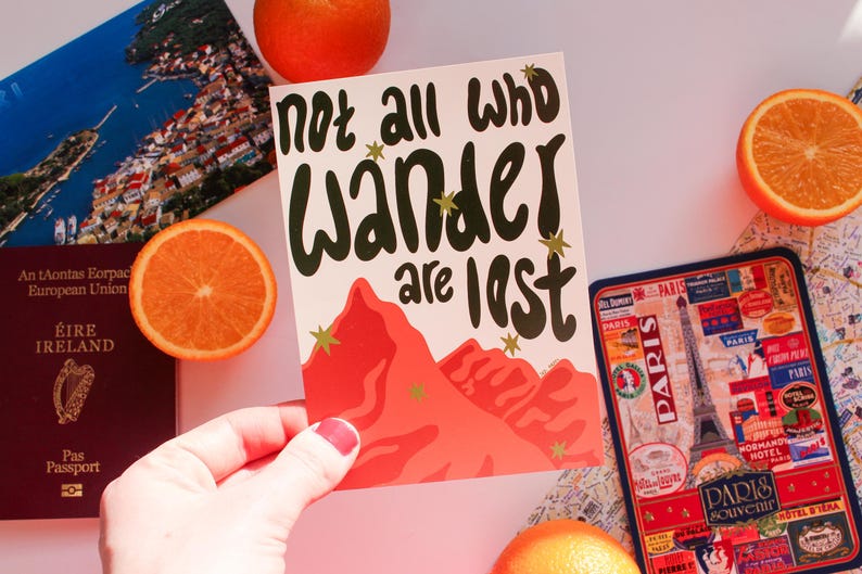 May include: A travel-themed image featuring a postcard with the text "not all who wander are lost" and a mountain illustration. Other items include a passport, a Paris souvenir, and orange slices. The postcard is held by a person.