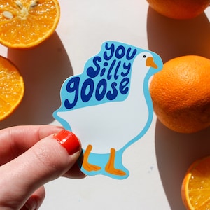 May include: A white goose sticker with blue text that says "You Silly Goose". The sticker is being held by a hand with red nail polish. There are oranges in the background.
