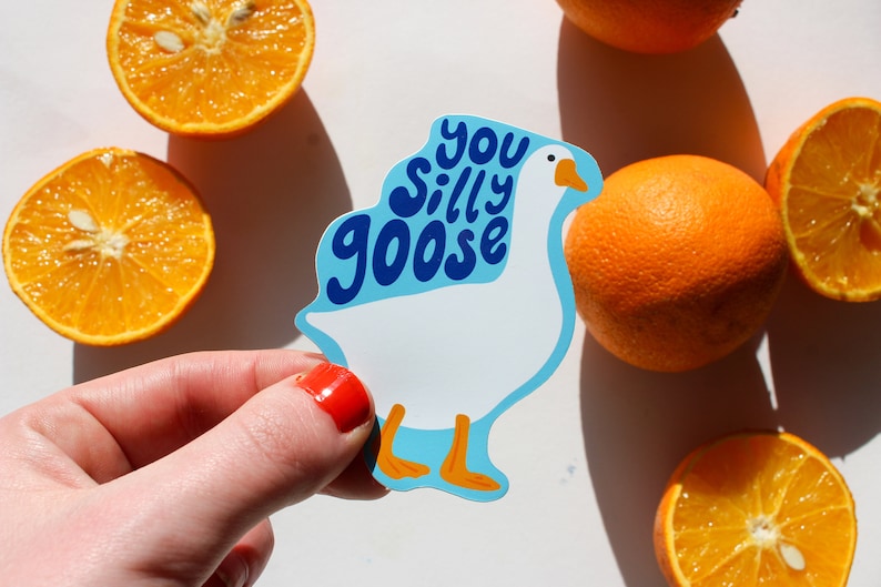 May include: A white goose sticker with blue text that says "You Silly Goose". The sticker is being held by a hand with red nail polish. There are oranges in the background.