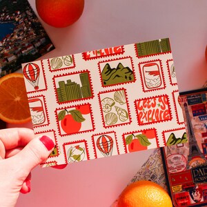 May include: A postcard with a stamp design featuring travel-themed illustrations, including a hot air balloon, cityscapes, and fruit. The postcard has the text "Let's Go Explore" and is held near oranges, a passport, and a travel-themed phone case.