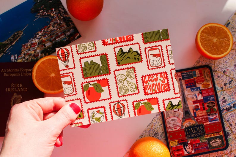 May include: A postcard with a stamp design featuring travel-themed illustrations, including a hot air balloon, cityscapes, and fruit. The postcard has the text "Let's Go Explore" and is held near oranges, a passport, and a travel-themed phone case.