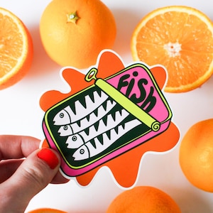 May include: A sticker depicting a pink sardine tin with the word "FISH" in white, set against an orange splash. The sticker shows three fish illustrations. Oranges and orange slices are arranged around the sticker.
