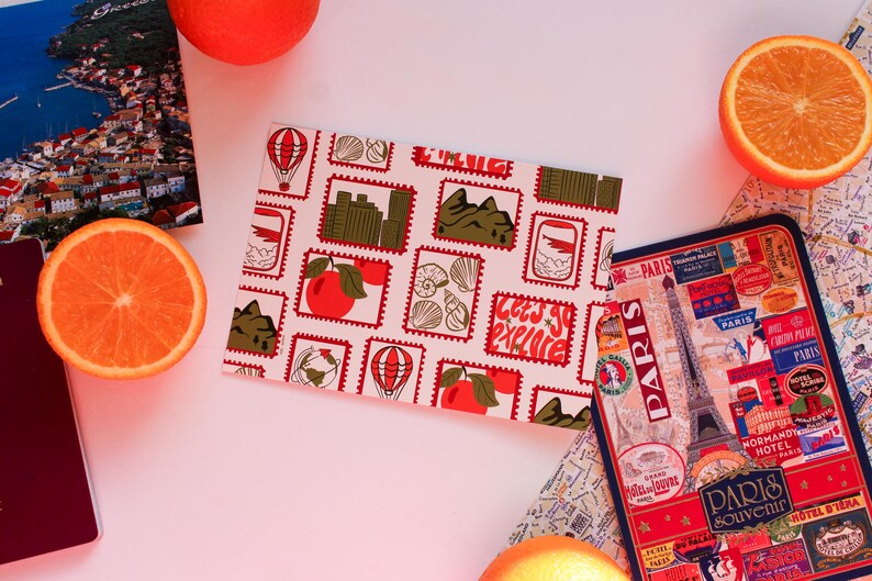 May include: A travel-themed flat lay featuring a patterned passport cover with stamps, a Paris souvenir, a map, and orange slices. The passport cover has the text "Let's go explore".