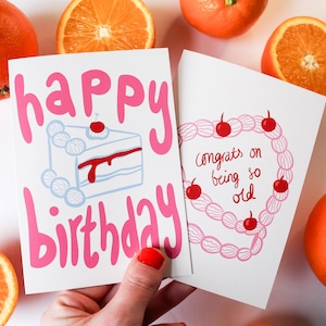 May include: Two pink and white birthday cards with illustrations. The first card says "happy birthday" with a pink cake and red cherries. The second card says "Congrats on being so old" with a pink string of cherries.