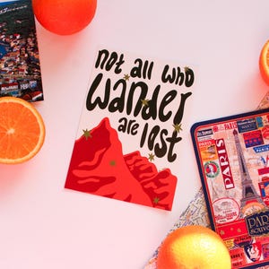 May include: A travel-themed still life featuring a postcard with the text "Not all who wander are lost", a passport, a Paris souvenir tray, a map, and orange slices. The postcard has a red mountain design.