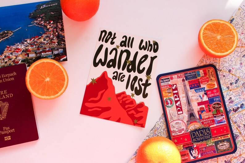 May include: A travel-themed still life featuring a postcard with the text "Not all who wander are lost", a passport, a Paris souvenir tray, a map, and orange slices. The postcard has a red mountain design.