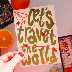 May include: A cream-colored card with the words "Let's Travel the World" in gold script, accented with red stars and a paper airplane. The image includes travel-themed items like a passport, a Paris souvenir, and orange slices.