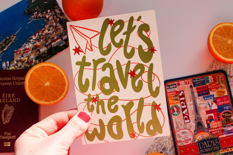 May include: A cream-colored card with the words "Let's Travel the World" in gold script, accented with red stars and a paper airplane. The image includes travel-themed items like a passport, a Paris souvenir, and orange slices.