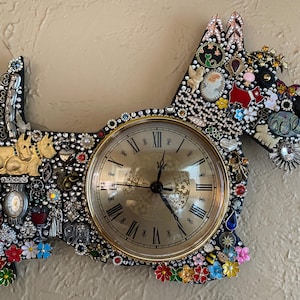 May include: A decorative wall clock in the shape of a dog, adorned with various embellishments. The clock face has Roman numerals and a gold-colored frame. The dog's body is covered in rhinestones, brooches, and colorful floral accents.