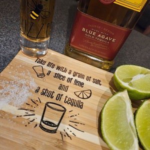 May include: A wooden cutting board with the text "Take life with a grain of salt, a slice of lime and a shot of tequila" engraved on it. There is a shot glass of tequila, a bottle of tequila, and lime wedges on the board.