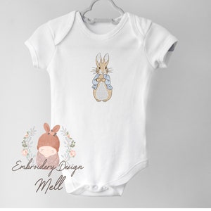 May include: White baby onesie with an embroidered image of Peter Rabbit in a blue jacket. The onesie has short sleeves and a snap closure. The image also includes the text "Embroidery Design Mell".