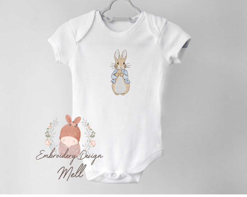 May include: White baby onesie with an embroidered image of Peter Rabbit in a blue jacket. The onesie has short sleeves and a snap closure. The image also includes the text "Embroidery Design Mell".