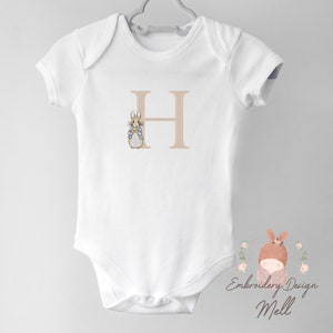 May include: White baby onesie with a beige embroidered letter H and a beige embroidered Peter Rabbit illustration. The onesie has a snap closure at the bottom. The text "Embroidery Design Mell" is printed in brown at the bottom of the onesie.