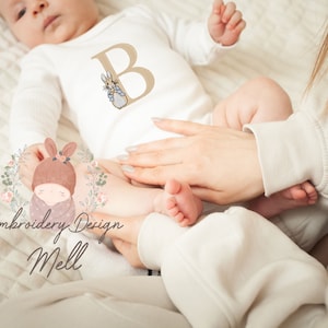 May include: A white baby onesie with a brown embroidered letter B and a small rabbit illustration. The onesie is worn by a baby lying on a white blanket. The text "Embroidery Design Mell" is visible in the lower left corner of the image.