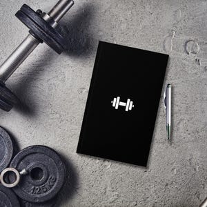 May include: A black notebook with a white dumbbell icon on the cover. A silver pen is lying next to the notebook. The notebook is on a gray concrete surface.