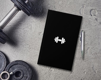 May include: A black notebook with a white dumbbell icon on the cover. A silver pen is lying next to the notebook. The notebook is on a gray concrete surface.