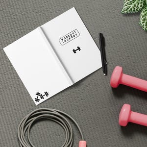 May include: A white workout logbook with a dumbbell icon and the text "WORKOUT LOGBOOK" on the cover. The logbook is lying on a gray surface with a jump rope, a pink dumbbell, and a black pen.