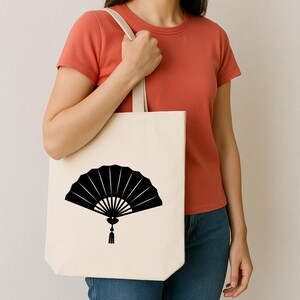 May include: A cream-colored tote bag featuring a black fan design. The fan is depicted in a stylized, open form with radiating lines and a tassel. The bag has a simple, minimalist design with a neutral color palette.