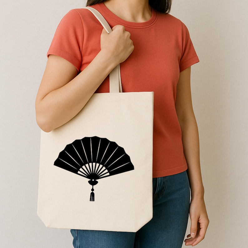 May include: A cream-colored tote bag featuring a black fan design. The fan is depicted in a stylized, open form with radiating lines and a tassel. The bag has a simple, minimalist design with a neutral color palette.