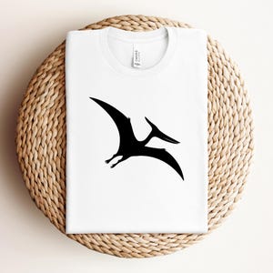 May include: White t-shirt with a black Pterodactyl silhouette. The design is centrally placed. The collar has the word "Canvas" printed on it. The shirt is displayed on a round, woven wicker surface.