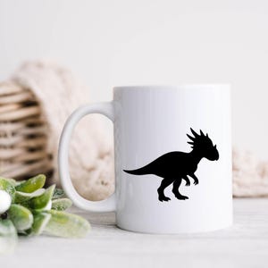 May include: White ceramic mug with a black silhouette of a Stygimoloch dinosaur. The dinosaur is shown in profile, with a spiky head and a long tail. The mug has a curved handle. The background is blurred.